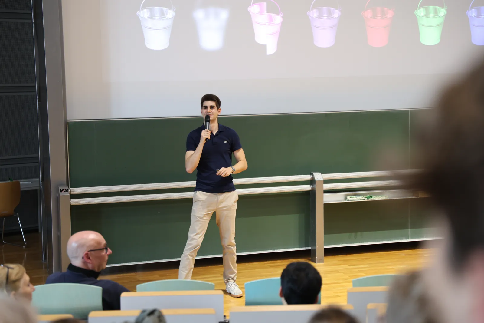 Dominik John on stage at the Science Slam Garching, with colourful paint buckets on the slide behind him